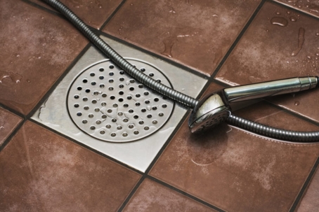 A shower head and hose are on a wet tiled floor, over a drain.