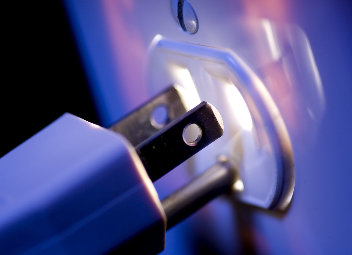 A plug inserted into a wall outlet. Metal prongs and white plastic.