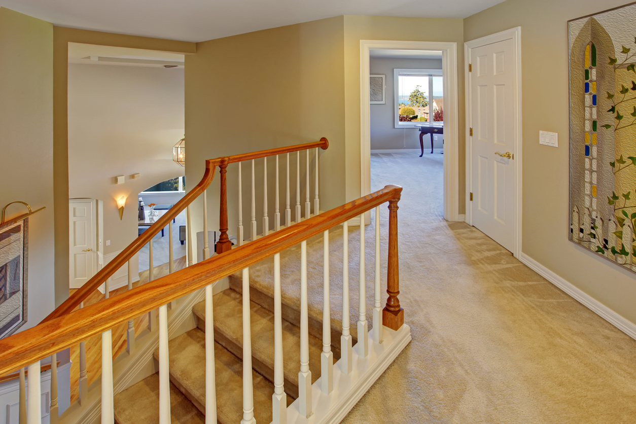 A light beige hallway with wooden stairs and railing, an open doorway to another room, and a colorful stained glass art piece on the wall.